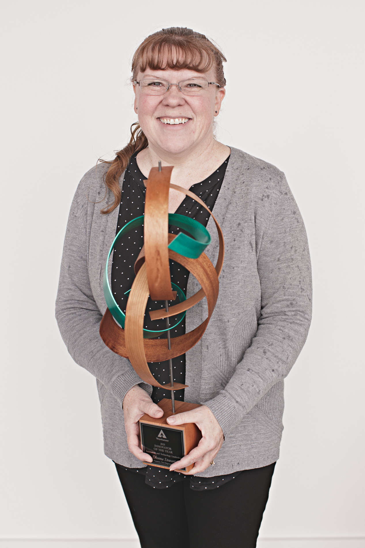 Image of Dionna Stewart. Dionna is wearing a gray cardigan sweather with her long-red hair over her shoulder. She wears glasses and holds the award, which is a twisted metal statue with a wood base.