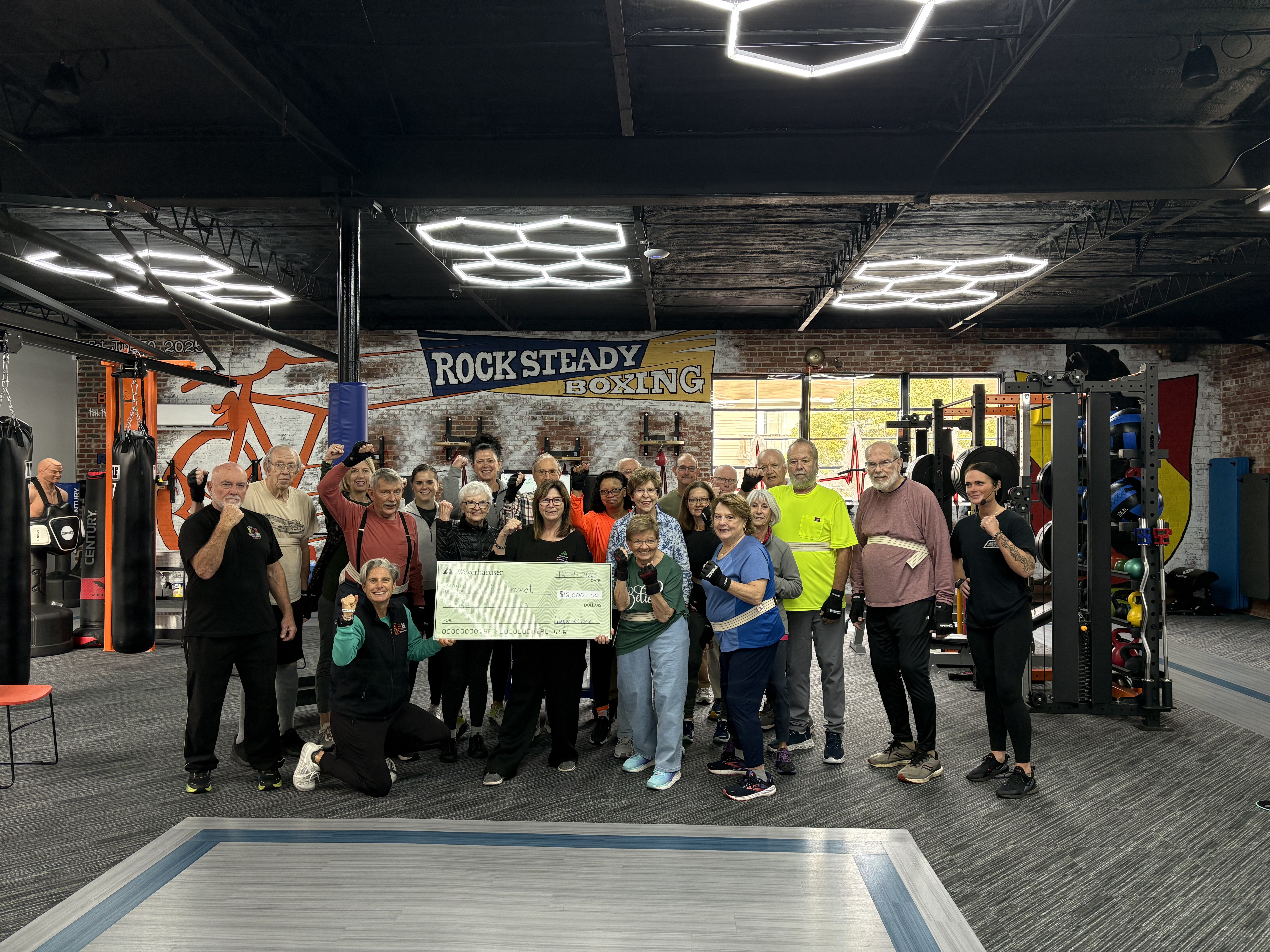 Image of Lori presenting a Giving Fund check to the Bike Box Project and Rock Steady Boxing. Lori is surriounded by numerous people who are training in a boxing gym.